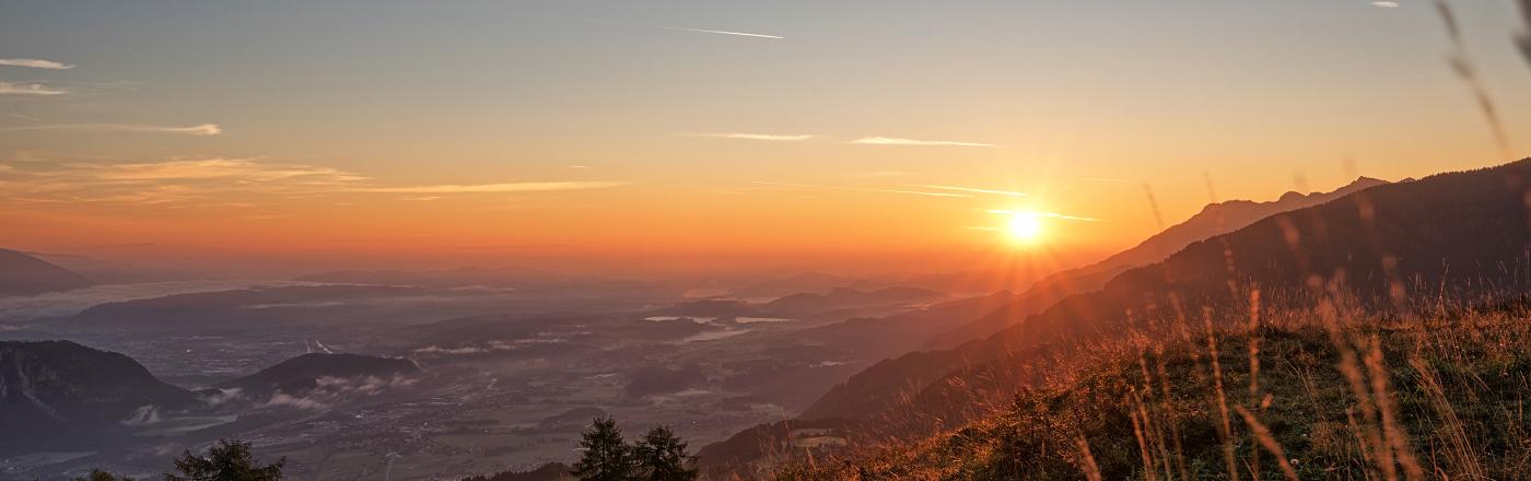 Ein atemberaubender Sonnenuntergang &uuml;ber einer malerischen Berglandschaft. Die orangefarbenen Strahlen der untergehenden Sonne tauchen den Himmel und das Tal in warmes Licht. Im Vordergrund sind Gr&auml;ser und B&auml;ume zu sehen, die eine friedliche Atmosph&auml;re schaffen.