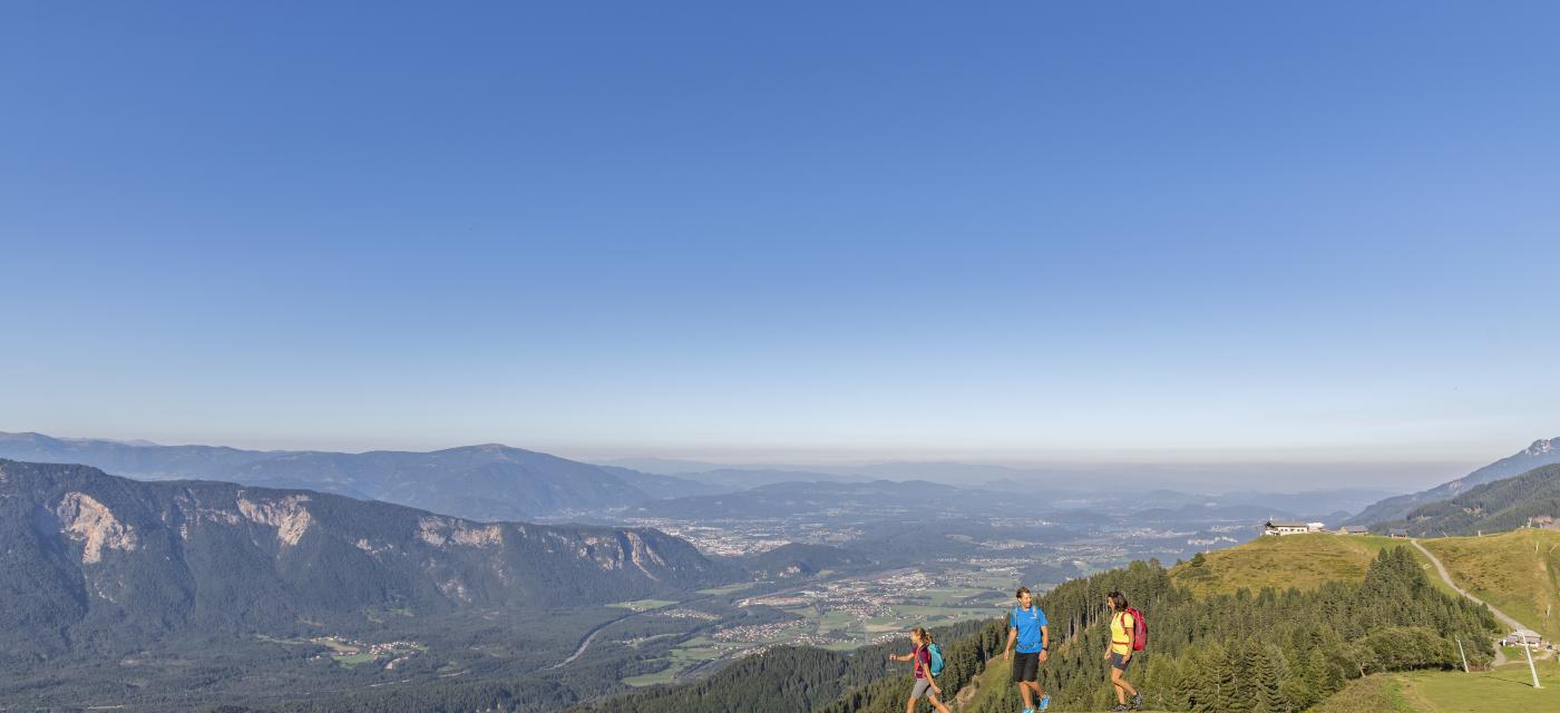 Drei Wanderer genießen einen sonnigen Tag in den Alpen. Vor ihnen breitet sich ein atemberaubender Blick über ein grünes Tal aus, während der klare blaue Himmel das Bild dominiert. Die Landschaft bietet eine perfekte Kulisse für Natur- und Outdoor-Liebhaber.