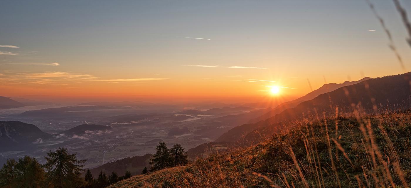 Ein atemberaubender Sonnenuntergang über einer malerischen Berglandschaft. Die orangefarbenen Strahlen der untergehenden Sonne tauchen den Himmel und das Tal in warmes Licht. Im Vordergrund sind Gräser und Bäume zu sehen, die eine friedliche Atmosphäre schaffen.
